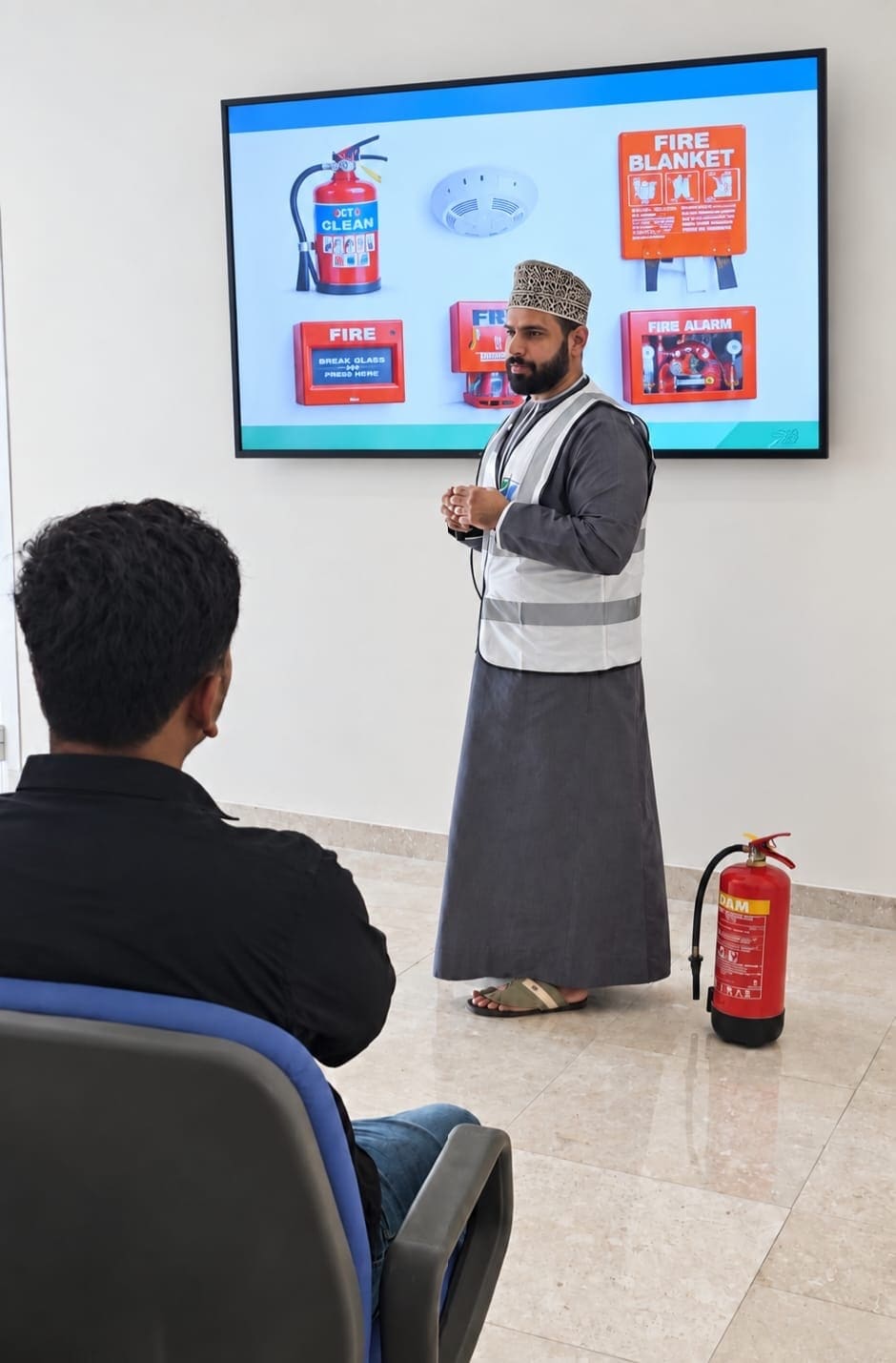 A KADI consultant in a high-visibility vest giving a fire safety session to MBBT staff, with extinguisher, alarm and fire blanket slides on the screen and an extinguisher on the floor beside him.