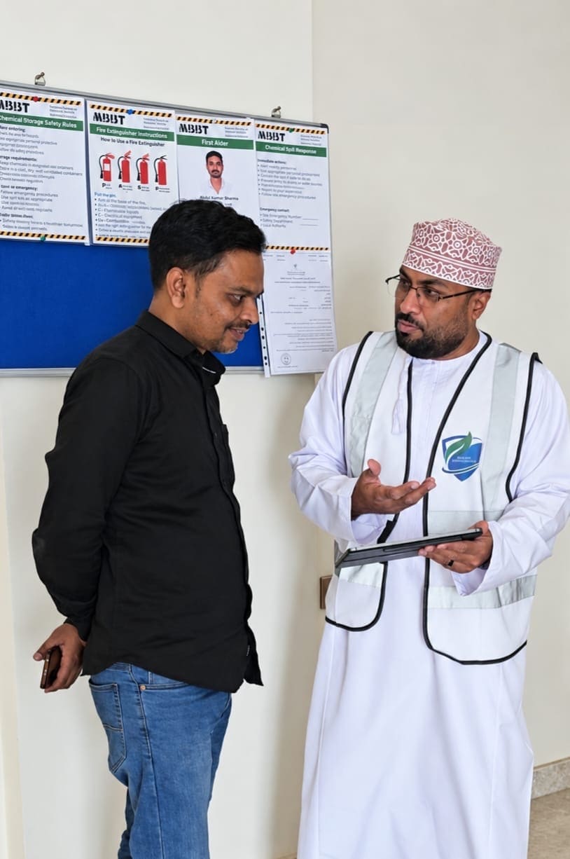 A KADI consultant in a high-visibility vest talking an MBBT team member through a point on a tablet beside the site's safety notice board.